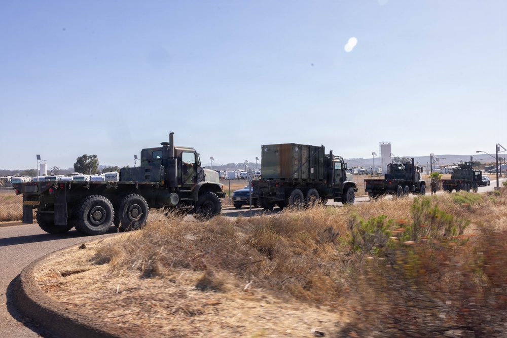 A convoy of three military trucks is traveling along a road in what appears to be a dry, arid environment consistent with a military base or installation. The vehicles are olive drab/camouflage colored heavy-duty tactical trucks, with one carrying a large enclosed cargo container. The surrounding terrain features dry grass and scrub vegetation under a clear blue sky.