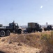 A convoy of three military trucks is traveling along a road in what appears to be a dry, arid environment consistent with a military base or installation. The vehicles are olive drab/camouflage colored heavy-duty tactical trucks, with one carrying a large enclosed cargo container. The surrounding terrain features dry grass and scrub vegetation under a clear blue sky.