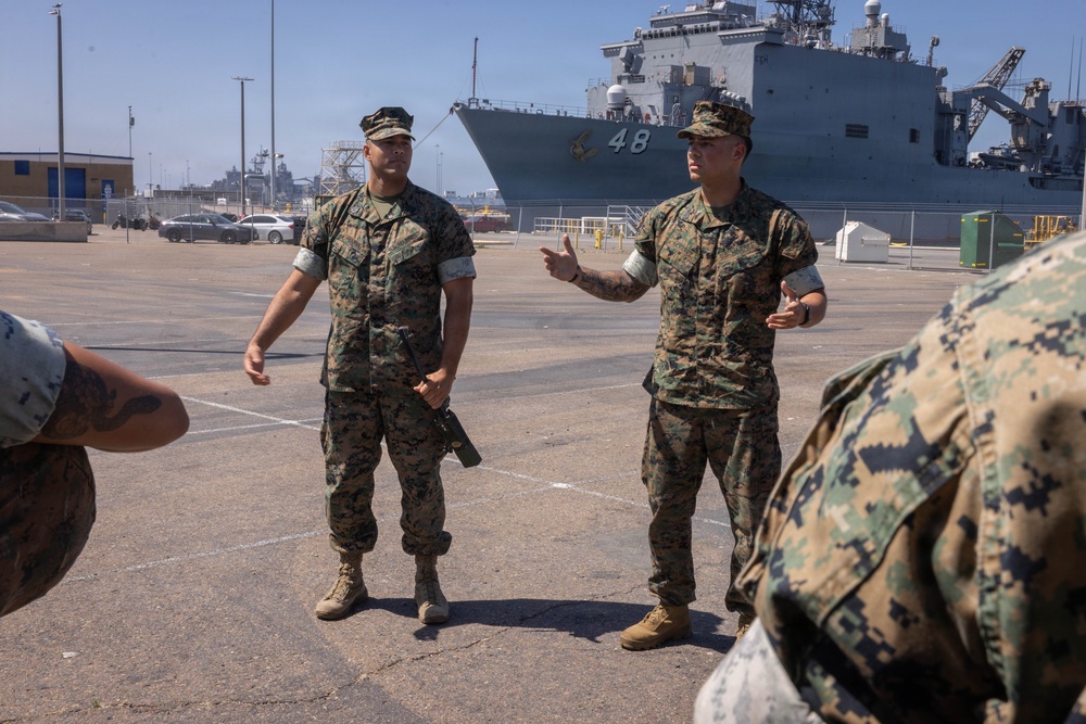 Two U.S. Marines in MARPAT camouflage uniforms stand on a naval pier, with one appearing to brief or instruct others. A large U.S. Navy amphibious ship bearing the hull number '48' is docked prominently in the background. The setting appears to be an active naval installation, likely on the U.S. West Coast.