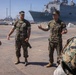 Two U.S. Marines in MARPAT camouflage uniforms stand on a naval pier, with one appearing to brief or instruct others. A large U.S. Navy amphibious ship bearing the hull number '48' is docked prominently in the background. The setting appears to be an active naval installation, likely on the U.S. West Coast.