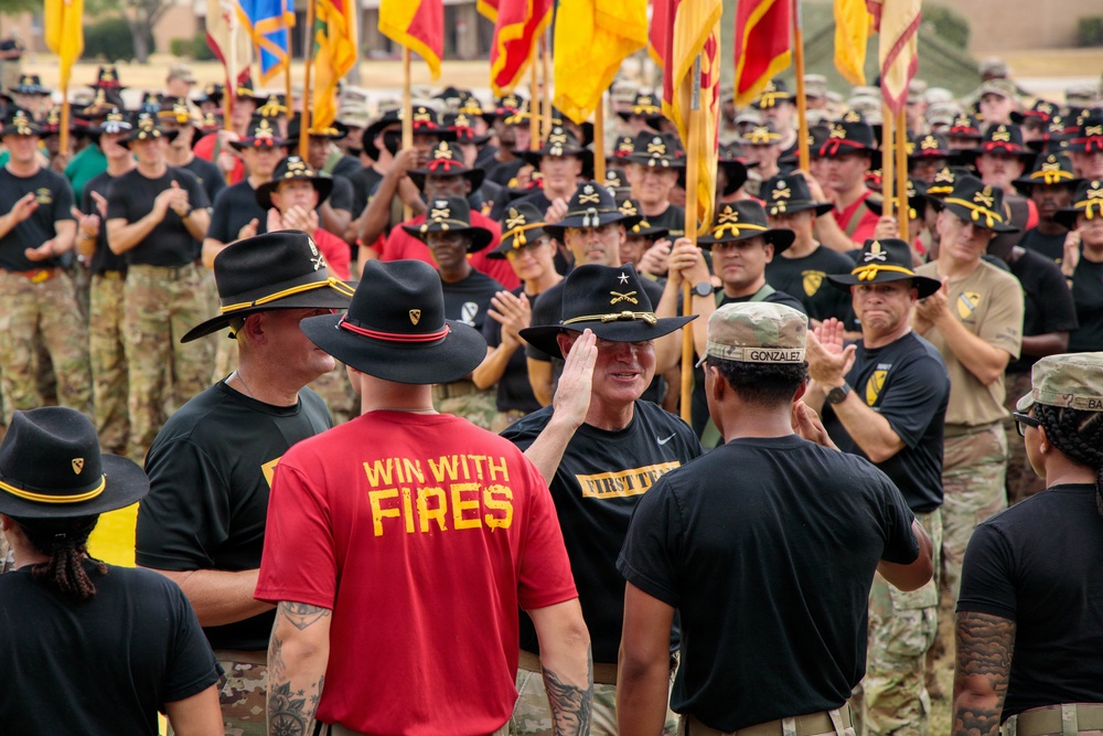 A large military formation event featuring U.S. Army soldiers, likely from the 1st Cavalry Division based on visible insignia and distinctive black campaign hats with crossed sabers. A senior officer (identifiable by a star on his hat) appears to be greeting or high-fiving a soldier named 'Gonzalez' in the foreground. Soldiers are wearing a mix of black unit PT shirts, red 'WIN WITH FIRES' shirts, and camouflage uniforms, with multiple unit guidon flags visible in the background.