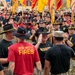 A large military formation event featuring U.S. Army soldiers, likely from the 1st Cavalry Division based on visible insignia and distinctive black campaign hats with crossed sabers. A senior officer (identifiable by a star on his hat) appears to be greeting or high-fiving a soldier named 'Gonzalez' in the foreground. Soldiers are wearing a mix of black unit PT shirts, red 'WIN WITH FIRES' shirts, and camouflage uniforms, with multiple unit guidon flags visible in the background.