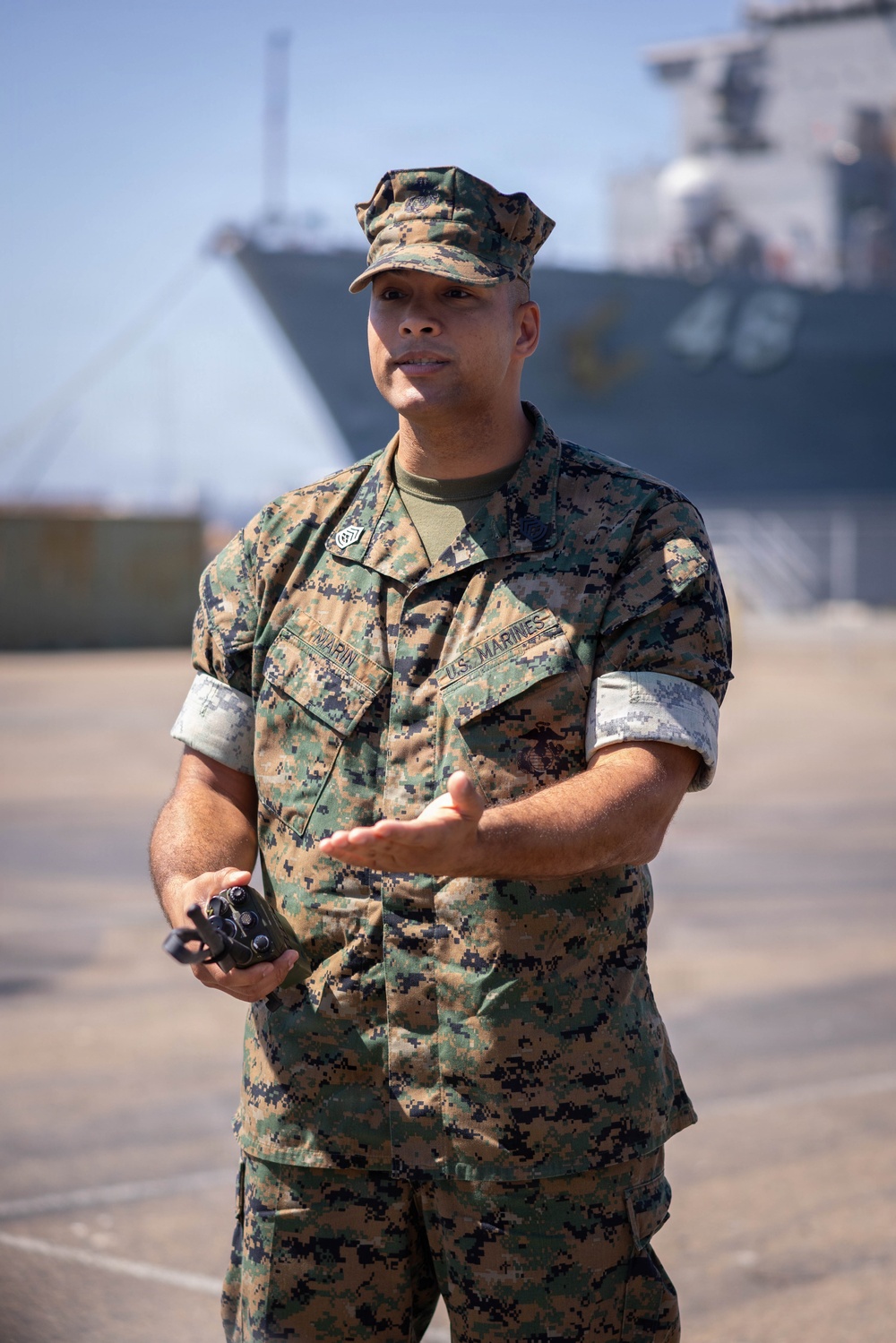 A U.S. Marine in MARPAT woodland camouflage uniform stands on what appears to be a pier or dock area, gesturing with one hand while holding a small device in the other. A large naval vessel is visible in the blurred background. The Marine's name tape reads 'MARIN' and the branch tape reads 'U.S. MARINES'.