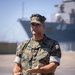 A U.S. Marine in MARPAT woodland camouflage uniform stands on what appears to be a pier or dock area, gesturing with one hand while holding a small device in the other. A large naval vessel is visible in the blurred background. The Marine's name tape reads 'MARIN' and the branch tape reads 'U.S. MARINES'.