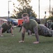 A large group of military personnel in olive green t-shirts and camouflage trousers are performing push-ups on a grass field, likely during a physical training session. The foreground features a tattooed male service member looking directly at the camera while in the up position of a push-up. The setting appears to be an outdoor military installation or base, photographed at dusk or dawn.