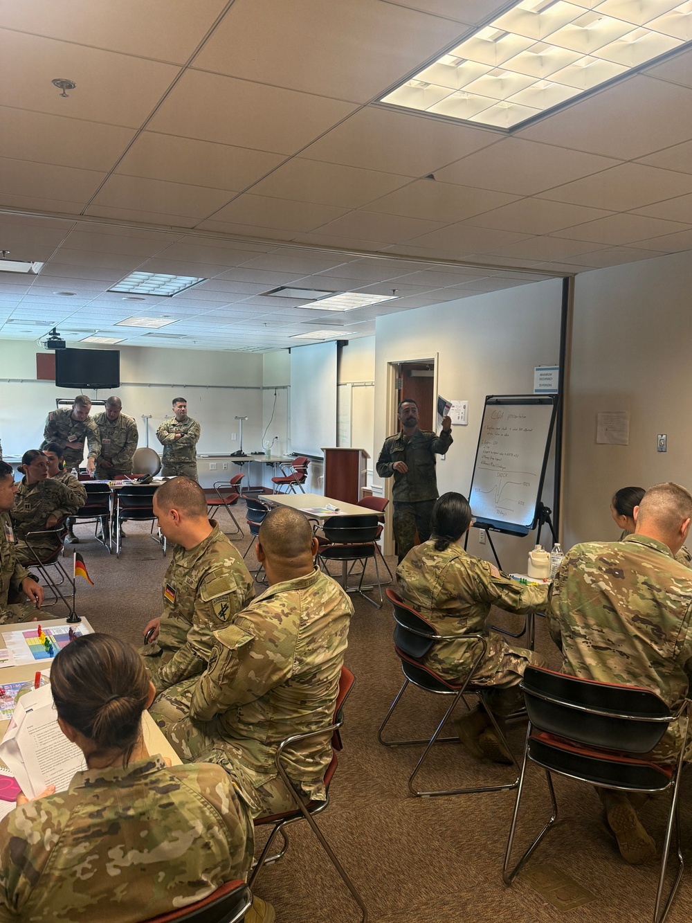 A military classroom or training session is underway in an indoor conference-style room. Personnel in camouflage uniforms from at least two nations are seated at tables while one individual stands at a flip chart presenting information. The setting appears to be a joint multinational military training or educational event.