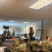 A military classroom or training session is underway in an indoor conference-style room. Personnel in camouflage uniforms from at least two nations are seated at tables while one individual stands at a flip chart presenting information. The setting appears to be a joint multinational military training or educational event.