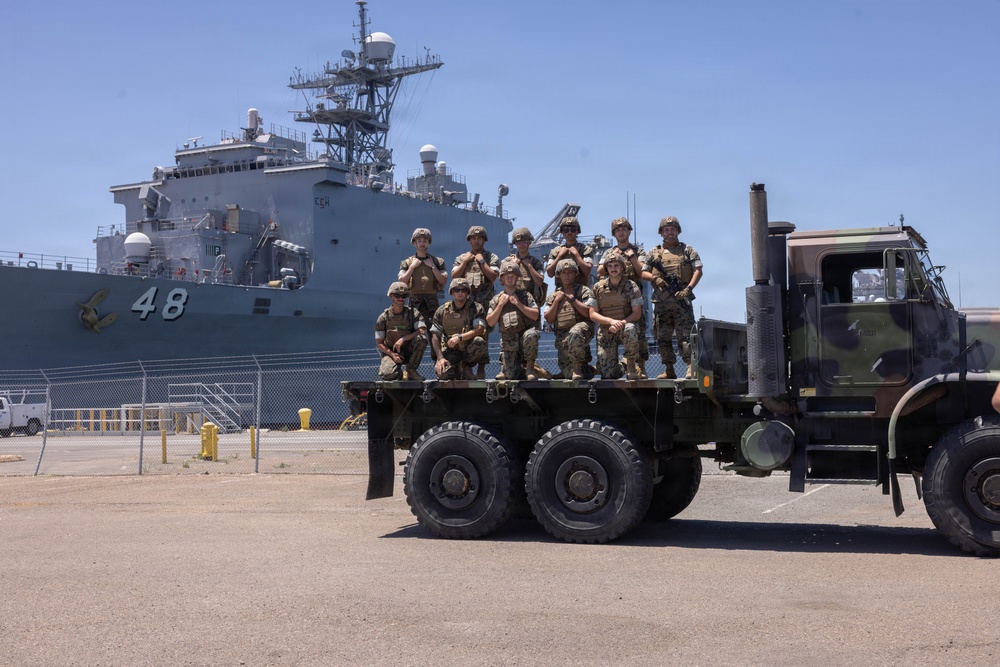 A group of approximately 10 military personnel in combat uniforms and tactical gear are posing for a group photo on the flatbed of a large military truck. They are positioned at a naval port facility with a large gray warship bearing the hull number '48' visible in the background. The setting appears to be a sunny day at a military harbor or naval base.