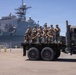 A group of approximately 10 military personnel in combat uniforms and tactical gear are posing for a group photo on the flatbed of a large military truck. They are positioned at a naval port facility with a large gray warship bearing the hull number '48' visible in the background. The setting appears to be a sunny day at a military harbor or naval base.