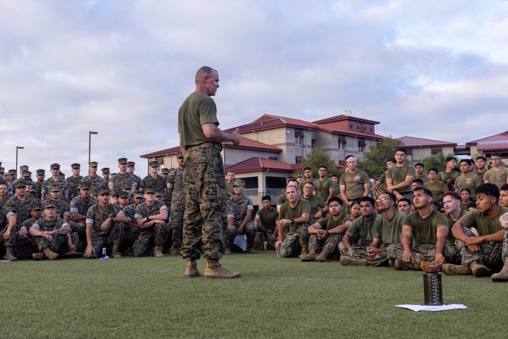 A senior military officer in camouflage utilities and olive drab t-shirt addresses a large formation of uniformed service members seated on a grass field. The gathering appears to be an outdoor briefing or all-hands formation at a military installation. The audience consists of dozens of personnel in matching camouflage uniforms attentively listening to the speaker.
