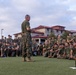 A senior military officer in camouflage utilities and olive drab t-shirt addresses a large formation of uniformed service members seated on a grass field. The gathering appears to be an outdoor briefing or all-hands formation at a military installation. The audience consists of dozens of personnel in matching camouflage uniforms attentively listening to the speaker.