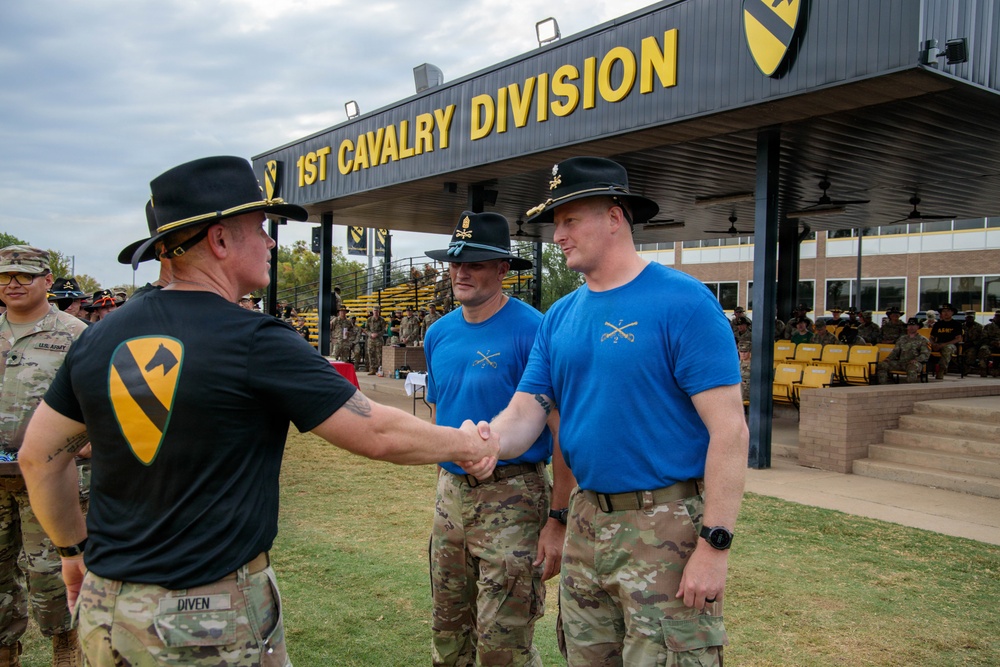 A military ceremony taking place at the 1st Cavalry Division headquarters, likely at Fort Cavazos (formerly Fort Hood), Texas. A soldier identified by nametape as 'DIVEN' wearing a black 1st Cavalry Division t-shirt and campaign hat is shaking hands with another soldier in a blue t-shirt bearing cavalry crossed sabers insignia. Two additional soldiers in matching blue shirts and campaign hats are present, suggesting a recognition or award ceremony.