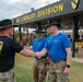 A military ceremony taking place at the 1st Cavalry Division headquarters, likely at Fort Cavazos (formerly Fort Hood), Texas. A soldier identified by nametape as 'DIVEN' wearing a black 1st Cavalry Division t-shirt and campaign hat is shaking hands with another soldier in a blue t-shirt bearing cavalry crossed sabers insignia. Two additional soldiers in matching blue shirts and campaign hats are present, suggesting a recognition or award ceremony.