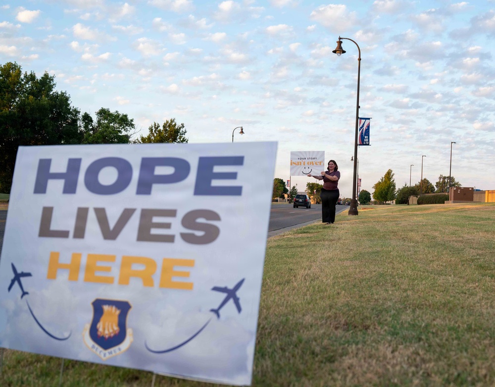 A person stands along a roadway on a military installation holding a sign that reads 'Your Story Isn't Over,' next to a prominent yard sign reading 'Hope Lives Here' featuring an Air Force emblem and aircraft silhouettes. The scene appears to be part of a suicide prevention or mental health awareness campaign. The setting is likely a U.S. Air Force base, suggested by the official insignia on the foreground sign.