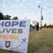 A person stands along a roadway on a military installation holding a sign that reads 'Your Story Isn't Over,' next to a prominent yard sign reading 'Hope Lives Here' featuring an Air Force emblem and aircraft silhouettes. The scene appears to be part of a suicide prevention or mental health awareness campaign. The setting is likely a U.S. Air Force base, suggested by the official insignia on the foreground sign.