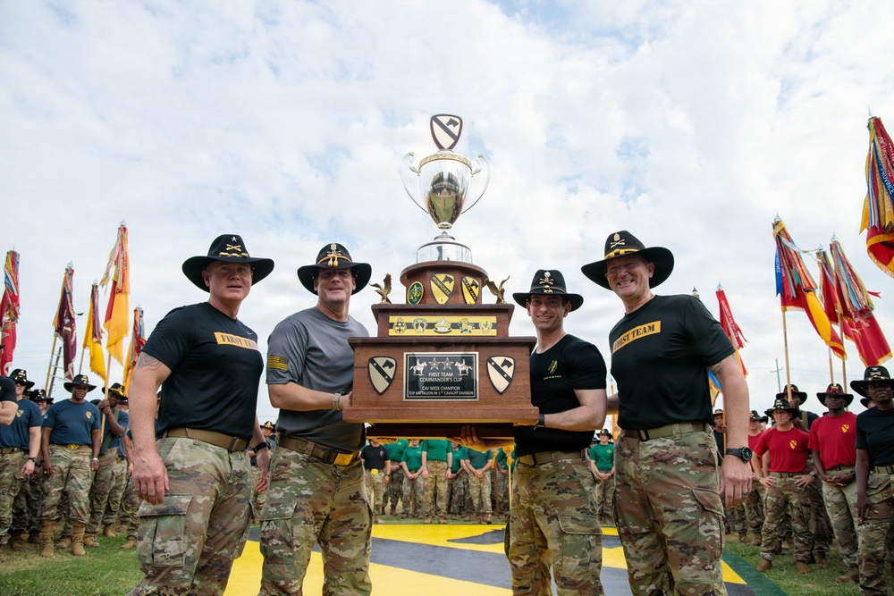 Four military personnel wearing campaign (cavalry) hats are holding a large trophy together at what appears to be an awards ceremony. The trophy is labeled 'First Team Commander's Cup' and features the 1st Cavalry Division insignia. The event is taking place outdoors with numerous soldiers and unit guidons/flags visible in the background.