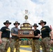 Four military personnel wearing campaign (cavalry) hats are holding a large trophy together at what appears to be an awards ceremony. The trophy is labeled 'First Team Commander's Cup' and features the 1st Cavalry Division insignia. The event is taking place outdoors with numerous soldiers and unit guidons/flags visible in the background.