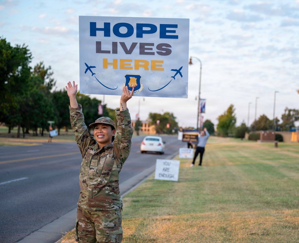 A U.S. Air Force service member in OCP (Operational Camouflage Pattern) uniform holds a 'Hope Lives Here' sign overhead along a military base roadway. The sign features the Air Force emblem and aircraft graphics, suggesting a mental health awareness or suicide prevention campaign. Additional signs and participants are visible in the background along the road.