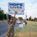 A U.S. Air Force service member in OCP (Operational Camouflage Pattern) uniform holds a 'Hope Lives Here' sign overhead along a military base roadway. The sign features the Air Force emblem and aircraft graphics, suggesting a mental health awareness or suicide prevention campaign. Additional signs and participants are visible in the background along the road.