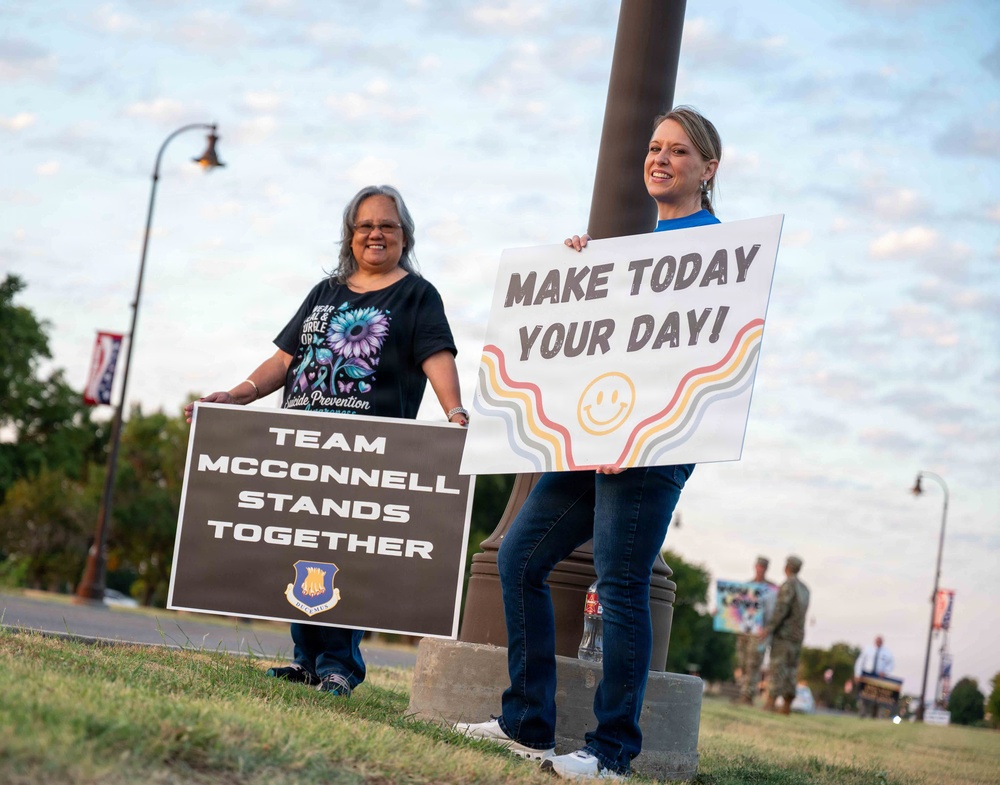 Two women stand outdoors on a grassy area near a lamppost, holding signs related to a suicide prevention awareness event at McConnell Air Force Base. The woman on the left holds a dark sign reading 'Team McConnell Stands Together' featuring a military shield emblem, while the woman on the right holds a cheerful sign reading 'Make Today Your Day!' decorated with colorful wavy lines and a smiley face. Military personnel are visible in the background.