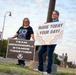 Two women stand outdoors on a grassy area near a lamppost, holding signs related to a suicide prevention awareness event at McConnell Air Force Base. The woman on the left holds a dark sign reading 'Team McConnell Stands Together' featuring a military shield emblem, while the woman on the right holds a cheerful sign reading 'Make Today Your Day!' decorated with colorful wavy lines and a smiley face. Military personnel are visible in the background.