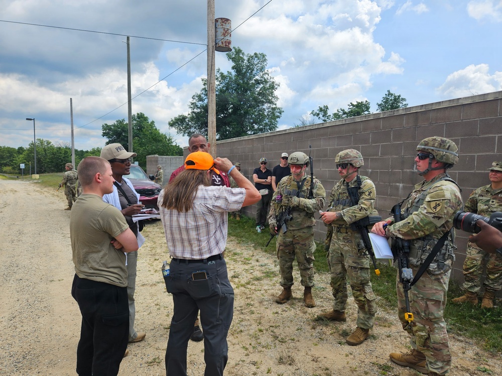 A group of military personnel in camouflage uniforms and combat gear are engaged in what appears to be a briefing or coordination meeting with several civilians near a concrete block wall along a dirt road. The mixed group includes both uniformed soldiers carrying rifles and civilians in casual attire, suggesting a joint exercise, training event, or site visit. The setting appears to be a military installation or training facility.
