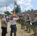 A group of military personnel in camouflage uniforms and combat gear are engaged in what appears to be a briefing or coordination meeting with several civilians near a concrete block wall along a dirt road. The mixed group includes both uniformed soldiers carrying rifles and civilians in casual attire, suggesting a joint exercise, training event, or site visit. The setting appears to be a military installation or training facility.