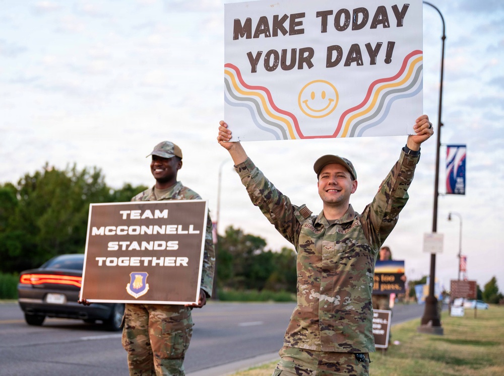 Two U.S. Air Force service members in OCP (Operational Camouflage Pattern) uniforms stand along a road holding motivational signs. The foreground airman raises a sign reading 'Make Today Your Day!' while the background airman holds a sign reading 'Team McConnell Stands Together,' referencing McConnell Air Force Base in Kansas. The scene appears to be a morale or suicide prevention awareness event.