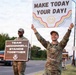 Two U.S. Air Force service members in OCP (Operational Camouflage Pattern) uniforms stand along a road holding motivational signs. The foreground airman raises a sign reading 'Make Today Your Day!' while the background airman holds a sign reading 'Team McConnell Stands Together,' referencing McConnell Air Force Base in Kansas. The scene appears to be a morale or suicide prevention awareness event.