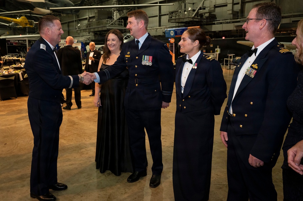 A formal military gala or dinner event taking place inside what appears to be an aviation museum or aircraft hangar. Military personnel in dress uniforms are greeting each other with handshakes, accompanied by civilian guests in formal attire. Several aircraft are visible in the background, confirming the museum/hangar setting.