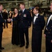 A formal military gala or dinner event taking place inside what appears to be an aviation museum or aircraft hangar. Military personnel in dress uniforms are greeting each other with handshakes, accompanied by civilian guests in formal attire. Several aircraft are visible in the background, confirming the museum/hangar setting.