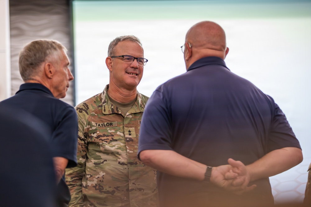 A U.S. Army officer wearing OCP (Operational Camouflage Pattern) uniform with the name tape 'TAYLOR' and two-star (Major General) rank insignia is engaged in conversation with two civilians in navy blue polo shirts. The setting appears to be an indoor venue, possibly a conference or briefing room with a large display screen in the background. The interaction appears informal and collegial.