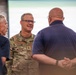 A U.S. Army officer wearing OCP (Operational Camouflage Pattern) uniform with the name tape 'TAYLOR' and two-star (Major General) rank insignia is engaged in conversation with two civilians in navy blue polo shirts. The setting appears to be an indoor venue, possibly a conference or briefing room with a large display screen in the background. The interaction appears informal and collegial.