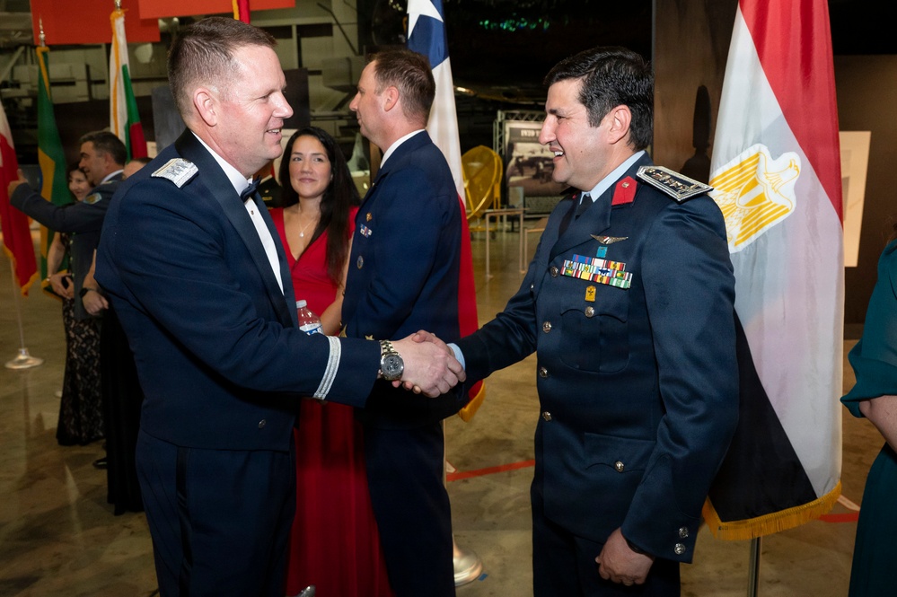 Two military officers in dress uniforms are shaking hands at what appears to be a formal military reception or gala event. One officer wears a US Air Force dress uniform while the other wears a darker uniform with numerous medals and insignia, standing near an Egyptian flag. Multiple national flags are displayed in the background, suggesting an international military gathering.