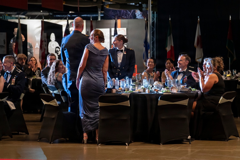 A formal military gala or dining-in event is taking place in what appears to be a large venue, possibly an aviation museum, with multiple national flags visible in the background. Military personnel in dress uniforms and civilians in formal attire are seated at round banquet tables with black linens. A couple appears to be approaching or being recognized at a central table while attendees applaud.