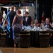 A formal military gala or dining-in event is taking place in what appears to be a large venue, possibly an aviation museum, with multiple national flags visible in the background. Military personnel in dress uniforms and civilians in formal attire are seated at round banquet tables with black linens. A couple appears to be approaching or being recognized at a central table while attendees applaud.