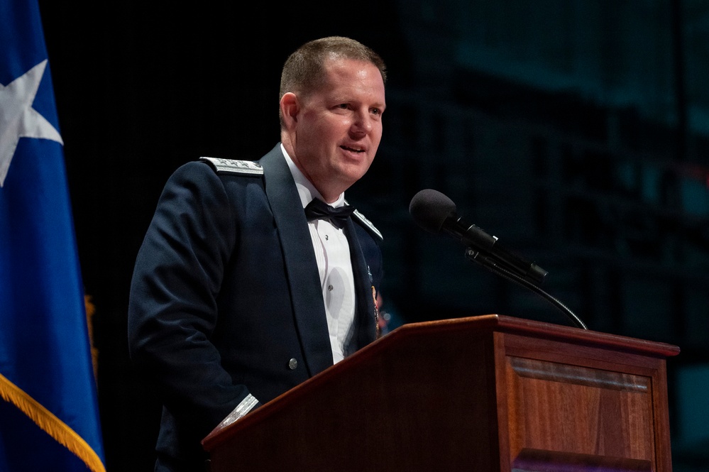 A military officer in dress uniform is speaking at a wooden podium with a microphone. He is wearing a dark blue formal military dress uniform with shoulder insignia indicating a general officer rank. A flag with a star is visible to his left, suggesting a formal military ceremony or banquet.