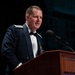 A military officer in dress uniform is speaking at a wooden podium with a microphone. He is wearing a dark blue formal military dress uniform with shoulder insignia indicating a general officer rank. A flag with a star is visible to his left, suggesting a formal military ceremony or banquet.