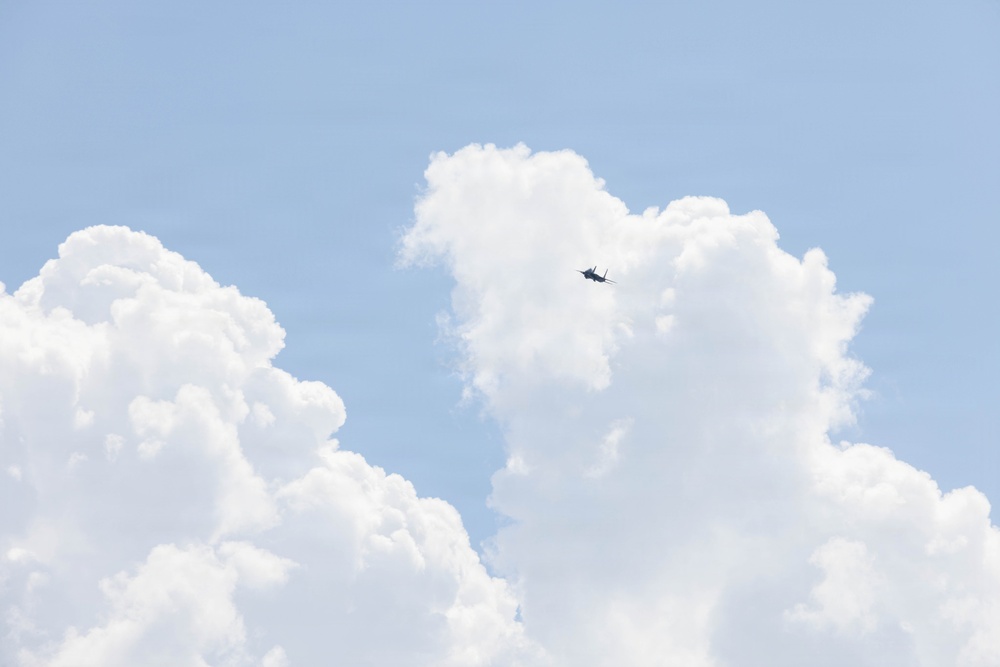A military jet aircraft is visible in flight against a blue sky with large cumulus clouds. The aircraft appears as a small dark silhouette positioned between cloud formations in the upper-center portion of the image. The scene suggests high-altitude flight in fair weather conditions.