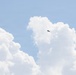 A military jet aircraft is visible in flight against a blue sky with large cumulus clouds. The aircraft appears as a small dark silhouette positioned between cloud formations in the upper-center portion of the image. The scene suggests high-altitude flight in fair weather conditions.