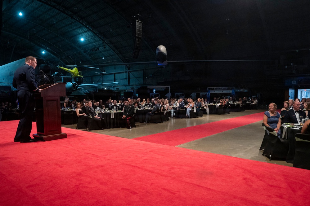 A formal military gala or ball is being held inside a large aviation museum or aircraft hangar. A uniformed speaker addresses a seated audience of military personnel and civilians at round banquet tables arranged along a red carpet aisle. Historic aircraft are visible in the background, serving as a dramatic backdrop for the event.