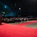 A formal military gala or ball is being held inside a large aviation museum or aircraft hangar. A uniformed speaker addresses a seated audience of military personnel and civilians at round banquet tables arranged along a red carpet aisle. Historic aircraft are visible in the background, serving as a dramatic backdrop for the event.