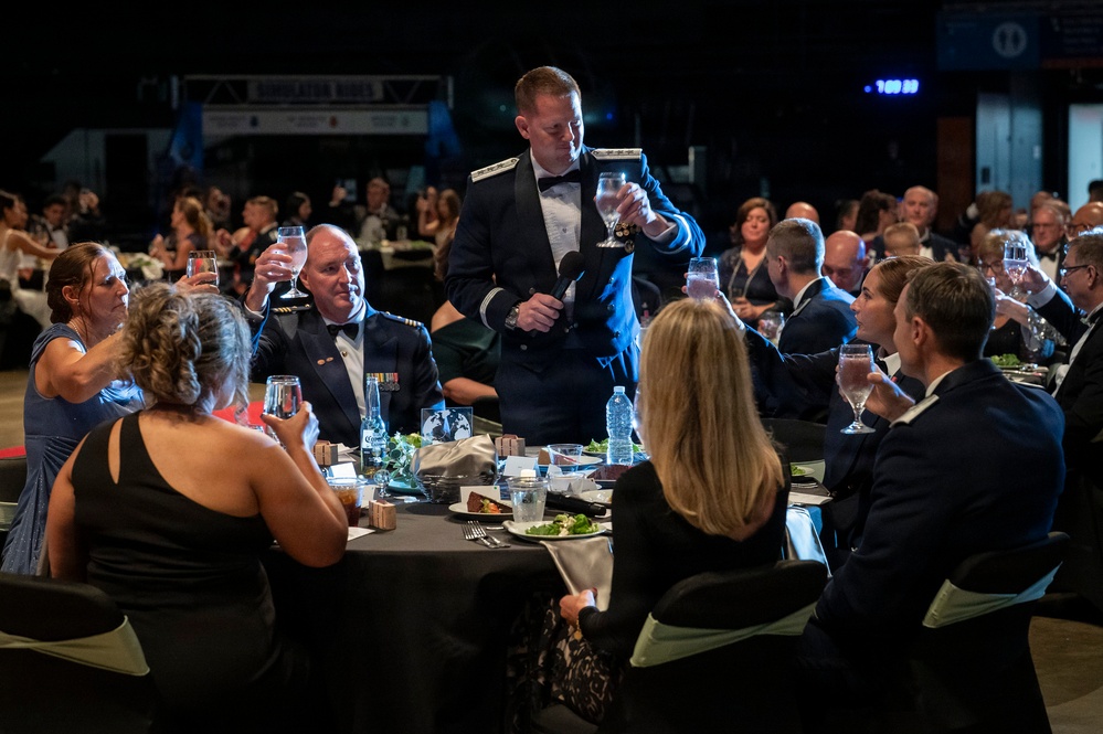 A formal military banquet or gala is taking place, with uniformed Air Force officers and civilians in formal attire seated at round tables. A standing officer in dress blues is leading a toast, raising a glass while holding a microphone. The event appears to be a large-scale military ball or awards dinner held in a convention-style venue.