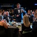 A formal military banquet or gala is taking place, with uniformed Air Force officers and civilians in formal attire seated at round tables. A standing officer in dress blues is leading a toast, raising a glass while holding a microphone. The event appears to be a large-scale military ball or awards dinner held in a convention-style venue.