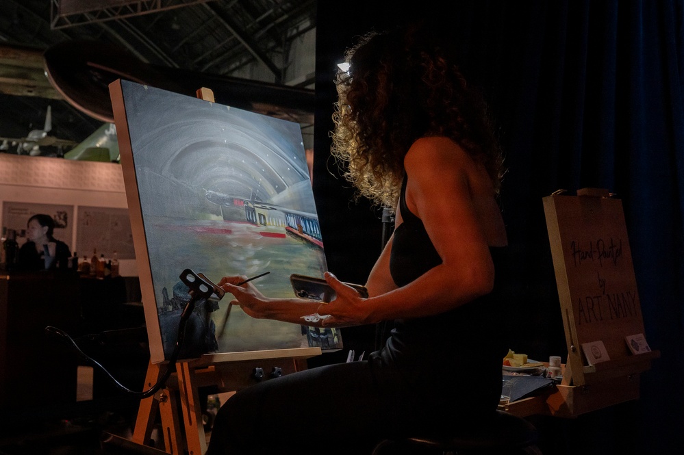A woman with curly hair is painting live at what appears to be an event or exhibition, working on a canvas depicting an aircraft inside a hangar or tunnel-like structure. She is seated at a wooden easel, holding a paintbrush and palette, illuminated by a small headlamp or spotlight. A sign to her right references 'Art Navy' or a similar organization, suggesting this is a live art demonstration event.