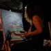 A woman with curly hair is painting live at what appears to be an event or exhibition, working on a canvas depicting an aircraft inside a hangar or tunnel-like structure. She is seated at a wooden easel, holding a paintbrush and palette, illuminated by a small headlamp or spotlight. A sign to her right references 'Art Navy' or a similar organization, suggesting this is a live art demonstration event.