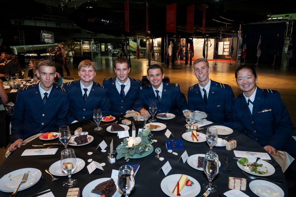 Six individuals in matching dark blue military-style uniforms are seated together at a formally set dining table, posing for a group photo. The setting appears to be a military aviation museum, with aircraft and exhibition displays visible in the background. The occasion appears to be a formal dinner or banquet event.