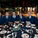 Six individuals in matching dark blue military-style uniforms are seated together at a formally set dining table, posing for a group photo. The setting appears to be a military aviation museum, with aircraft and exhibition displays visible in the background. The occasion appears to be a formal dinner or banquet event.