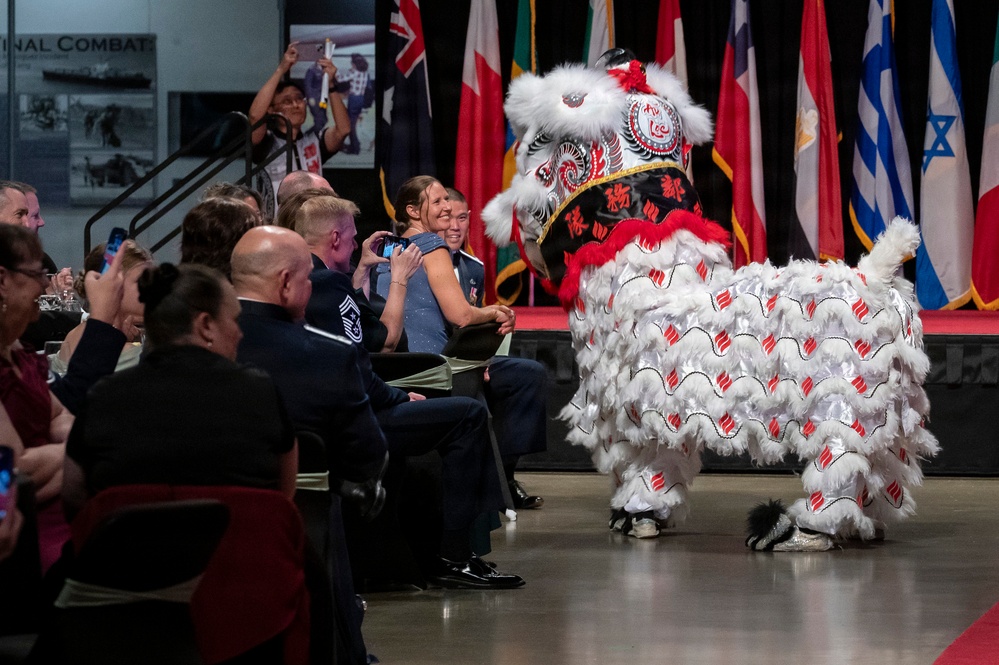 A traditional Chinese lion dance performance is taking place at what appears to be a formal military event or banquet, likely held at a museum or exhibition hall. The elaborate white, red, and black lion costume is interacting with seated audience members, many of whom are in military dress uniforms. International flags are displayed on a stage in the background.