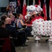 A traditional Chinese lion dance performance is taking place at what appears to be a formal military event or banquet, likely held at a museum or exhibition hall. The elaborate white, red, and black lion costume is interacting with seated audience members, many of whom are in military dress uniforms. International flags are displayed on a stage in the background.